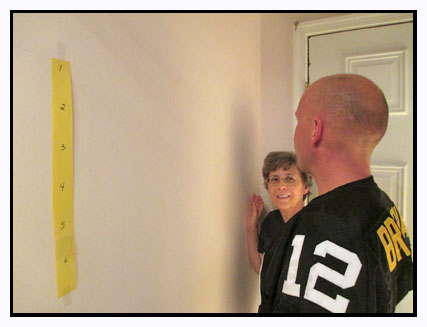 Photo shows the man in the football jersey looking toward the right on the wall in front of him.  There is a yellow strip on the wall to his left, but not where he is looking to the right.  Dona is watching him.
